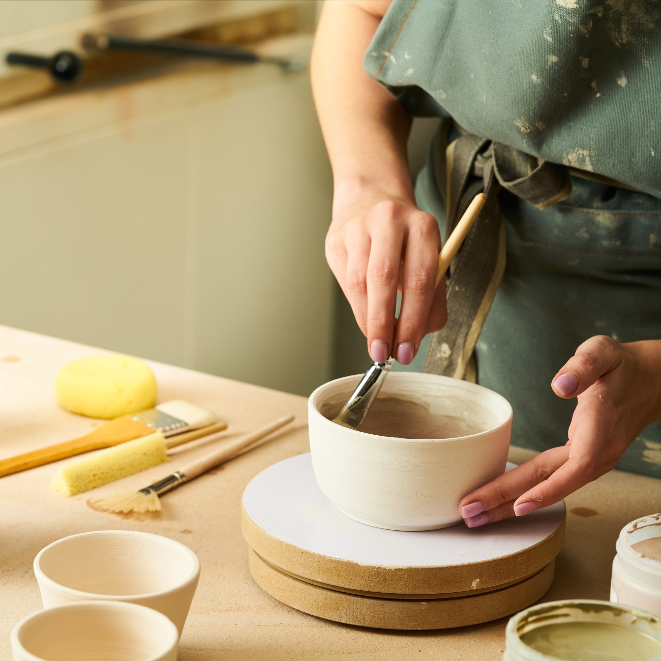 Close-up of girl painting clay mug with glaze. Woman coloring pottery in workshop with a paintbrush. Painter in green apron glazing clay pot.