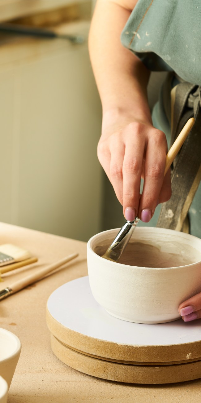 Close-up of girl painting clay mug with glaze. Woman coloring pottery in workshop with a paintbrush. Painter in green apron glazing clay pot.