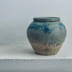 A handcrafted ceramic vessel by Sirin AlMasri with a rounded body and matching lid, featuring a textured stoneware base and layered blue glaze that transitions from rough clay to smooth finish, evoking geological formations and ancient pottery.