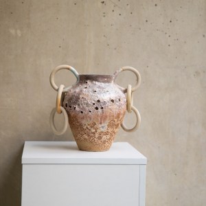 A ceramic vessel by Sirin AlMasri with a tall, amphora-inspired form, crater-textured glaze in earthy tones, and large ceramic rings attached to two looped side handles. The piece combines ancient symbolism with contemporary sculptural expression.
