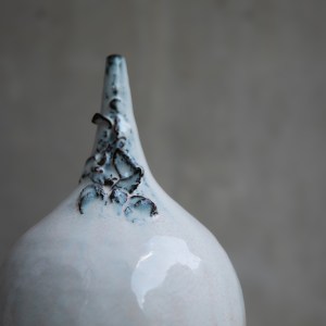 A ceramic vessel by Sirin AlMasri with a tall, amphora-inspired form, crater-textured glaze in earthy tones, and large ceramic rings attached to two looped side handles.