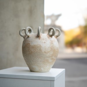 A sculptural ceramic vessel by Sirin AlMasri with a crater-textured surface in pale earth and rust tones, crowned with multiple looped protrusions around its rim. The form evokes ancient containers marked by time and elemental change.