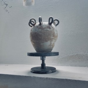 A sculptural ceramic vessel by Sirin AlMasri with a crater-textured surface in pale earth and rust tones, crowned with multiple looped protrusions around its rim. The form evokes ancient containers marked by time and elemental change.