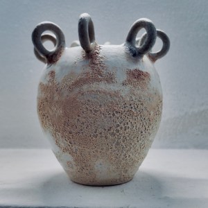 A sculptural ceramic vessel by Sirin AlMasri with a crater-textured surface in pale earth and rust tones, crowned with multiple looped protrusions around its rim. The form evokes ancient containers marked by time and elemental change.