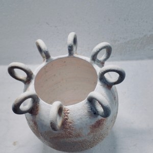 A sculptural ceramic vessel by Sirin AlMasri with a crater-textured surface in pale earth and rust tones, crowned with multiple looped protrusions around its rim. The form evokes ancient containers marked by time and elemental change.