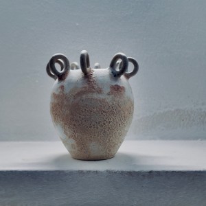 A sculptural ceramic vessel by Sirin AlMasri with a crater-textured surface in pale earth and rust tones, crowned with multiple looped protrusions around its rim. The form evokes ancient containers marked by time and elemental change.