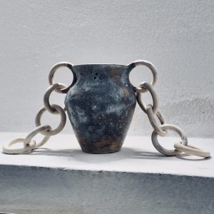 A ceramic vessel by Sirin AlMasri titled Nawah, with a cratered exterior, glossy turquoise interior, and sculptural ceramic chains on both sides. Inspired by the Earth's core, the piece explores density, tension, and elemental presence through clay and glaze.