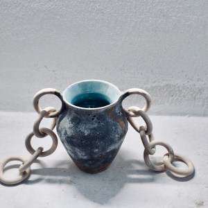 A ceramic vessel by Sirin AlMasri titled Nawah, with a cratered exterior, glossy turquoise interior, and sculptural ceramic chains on both sides. Inspired by the Earth's core, the piece explores density, tension, and elemental presence through clay and glaze.