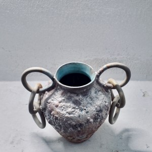 A ceramic vessel by Sirin AlMasri with a tall, amphora-inspired form, crater-textured glaze in earthy tones, and large ceramic rings attached to two looped side handles. The piece combines ancient symbolism with contemporary sculptural expression.