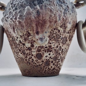 A ceramic vessel by Sirin AlMasri with a tall, amphora-inspired form, crater-textured glaze in earthy tones, and large ceramic rings attached to two looped side handles. The piece combines ancient symbolism with contemporary sculptural expression.