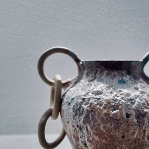 A ceramic vessel by Sirin AlMasri with a tall, amphora-inspired form, crater-textured glaze in earthy tones, and large ceramic rings attached to two looped side handles. The piece combines ancient symbolism with contemporary sculptural expression.