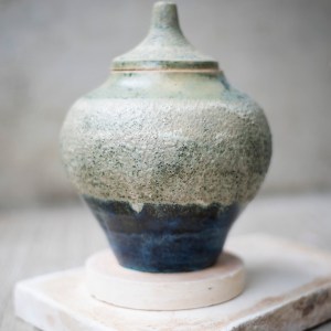 A lidded stoneware vessel by Sirin AlMasri with a conical top and rounded body, featuring a textured matte surface in mineral green and a deep, glossy blue glaze. The piece evokes natural elevation and contrasts between rough and smooth surfaces.