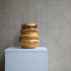 Handmade ceramic vase Rimal (رِمَال) by Zolta Studio, featuring a sculptural layered form with warm sandy brown glaze and natural dripping textures, displayed on a minimalist pedestal against a textured wall.