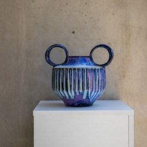 Handcrafted ceramic vase with carved vertical texture and twin circular handles, finished in flowing cobalt blue, violet, and indigo glazes. The sculptural form combines geometric lines with organic glaze movement, creating a striking contemporary ceramic artwork.