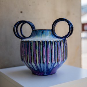Handcrafted ceramic vase with carved vertical texture and twin circular handles, finished in flowing cobalt blue, violet, and indigo glazes. The sculptural form combines geometric lines with organic glaze movement, creating a striking contemporary ceramic artwork.