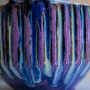 Handcrafted ceramic vase with carved vertical texture and twin circular handles, finished in flowing cobalt blue, violet, and indigo glazes. The sculptural form combines geometric lines with organic glaze movement, creating a striking contemporary ceramic artwork.