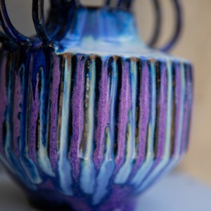 Handcrafted ceramic vase with carved vertical texture and twin circular handles, finished in flowing cobalt blue, violet, and indigo glazes. The sculptural form combines geometric lines with organic glaze movement, creating a striking contemporary ceramic artwork.